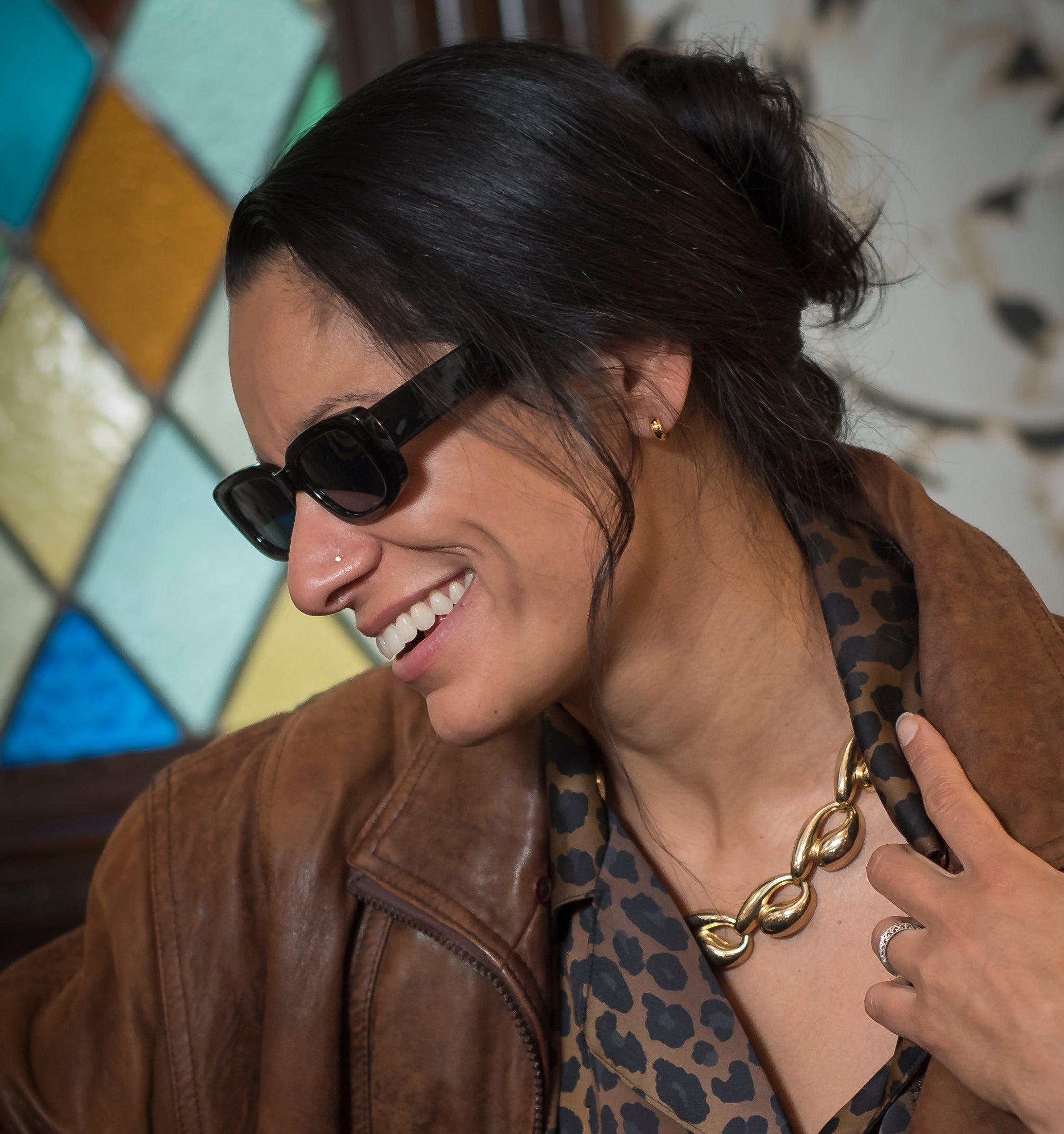 Woman wearing sunglasses and a brown jacket with a colorful stained glass window in the background