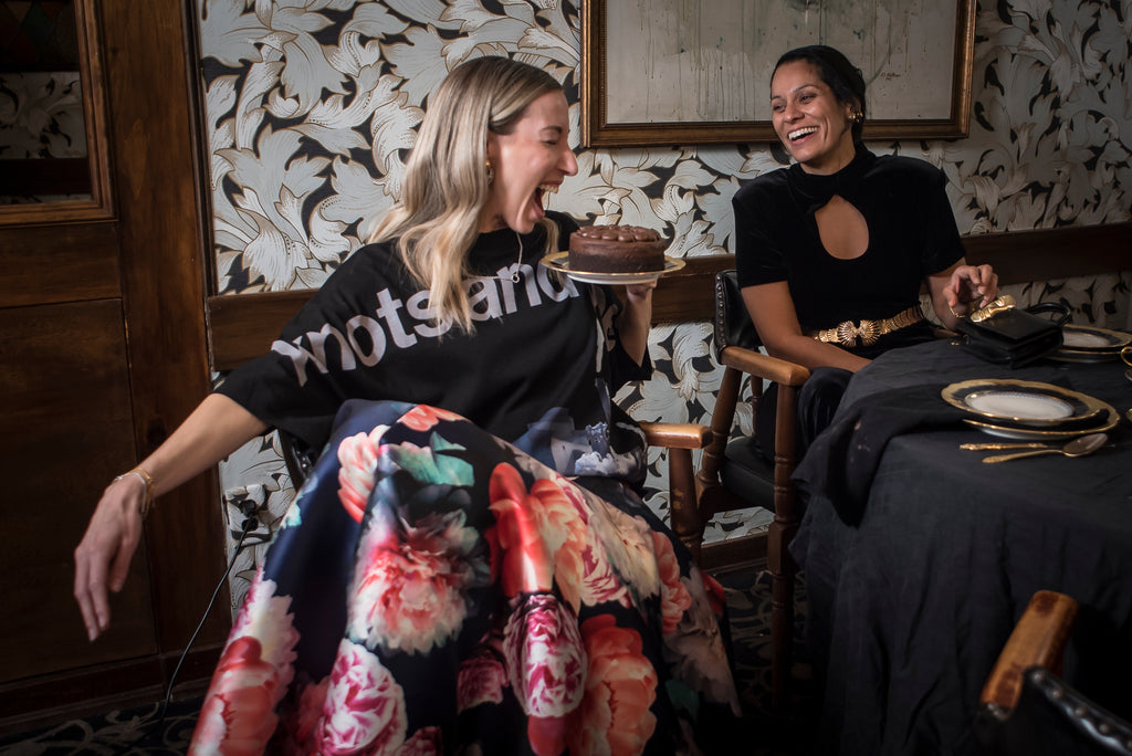 Two women sitting in a room with patterned wallpaper and a table with a cake.