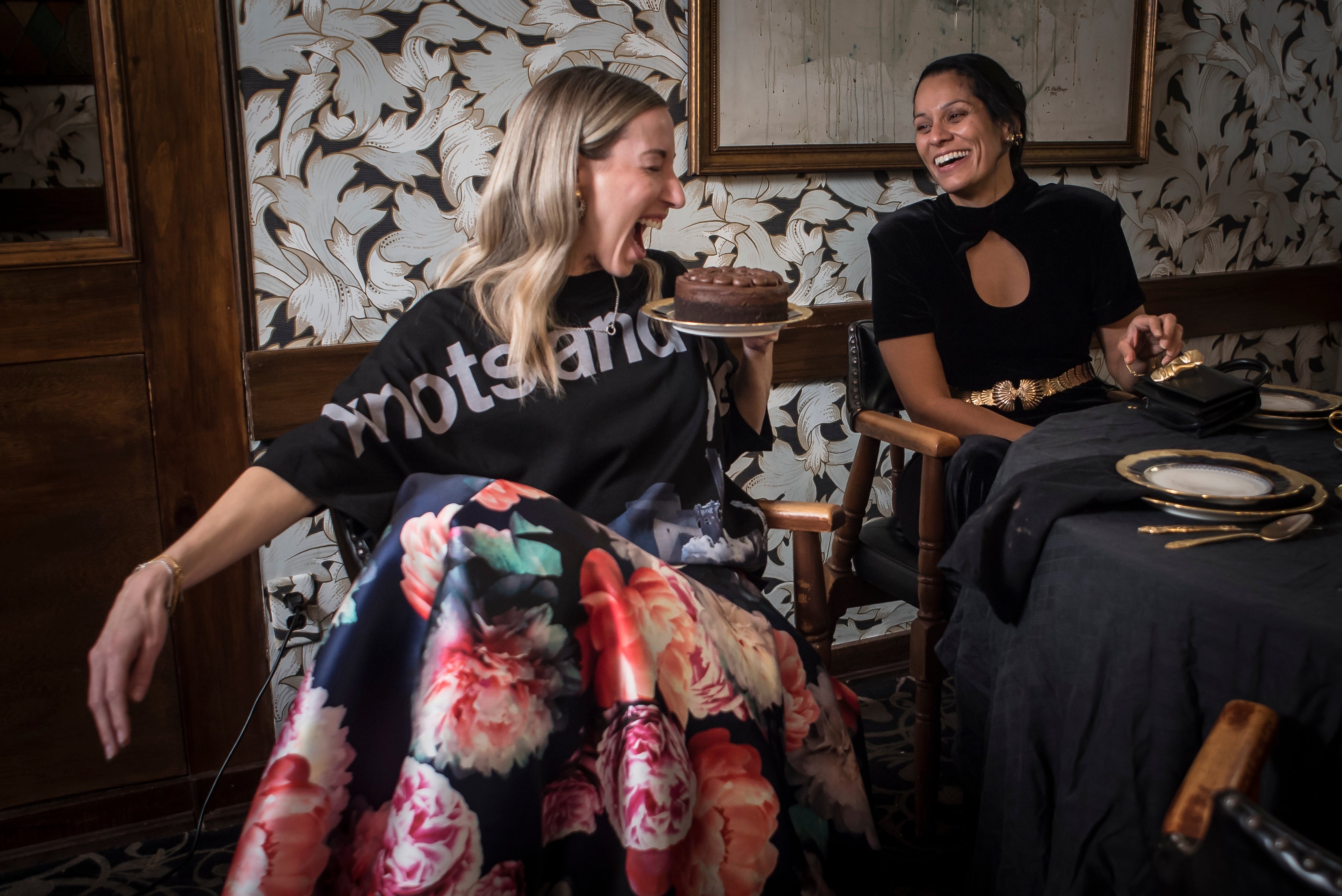 Two women sitting in a room with patterned wallpaper and a table with a cake.