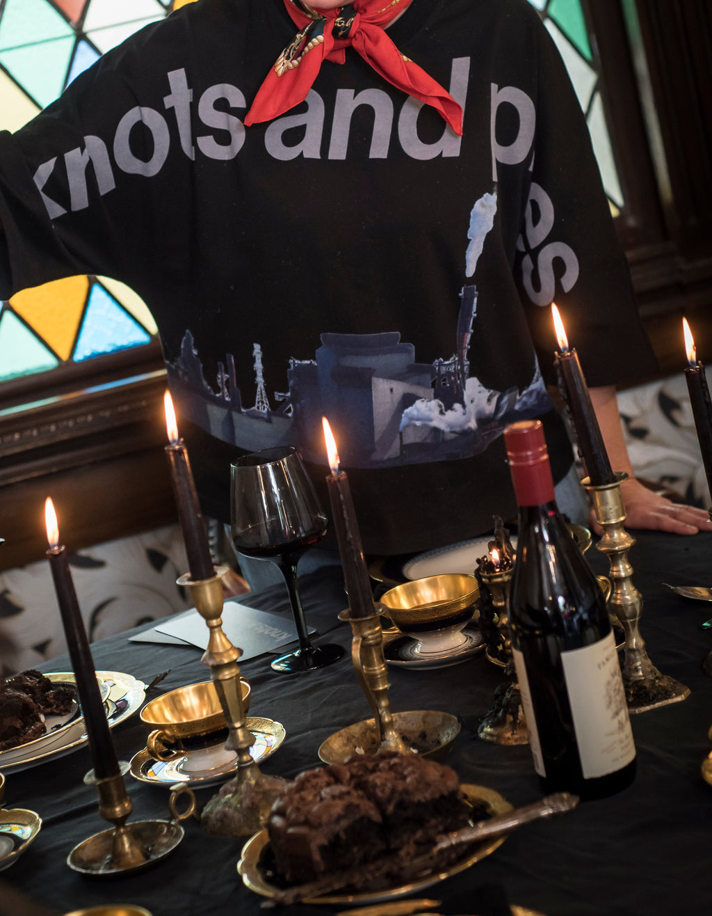 Person wearing a black shirt with text, standing behind a table with candles and bottles in a room with stained glass windows.
