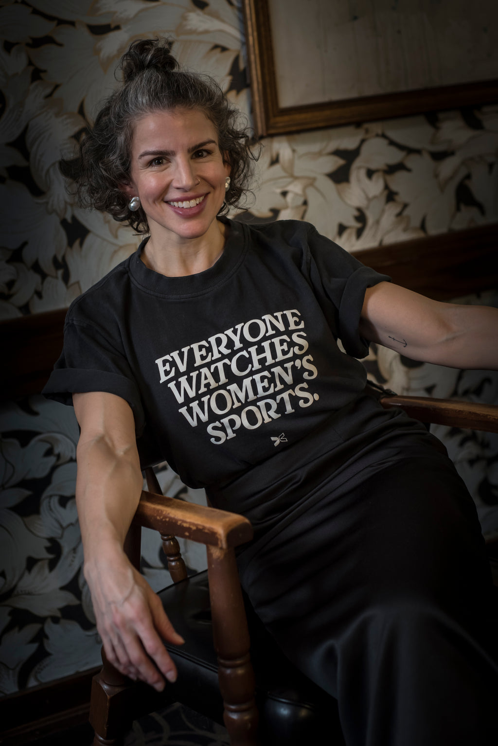Woman wearing a t-shirt with 'Everyone watches women's sports' text, sitting on a wooden chair.