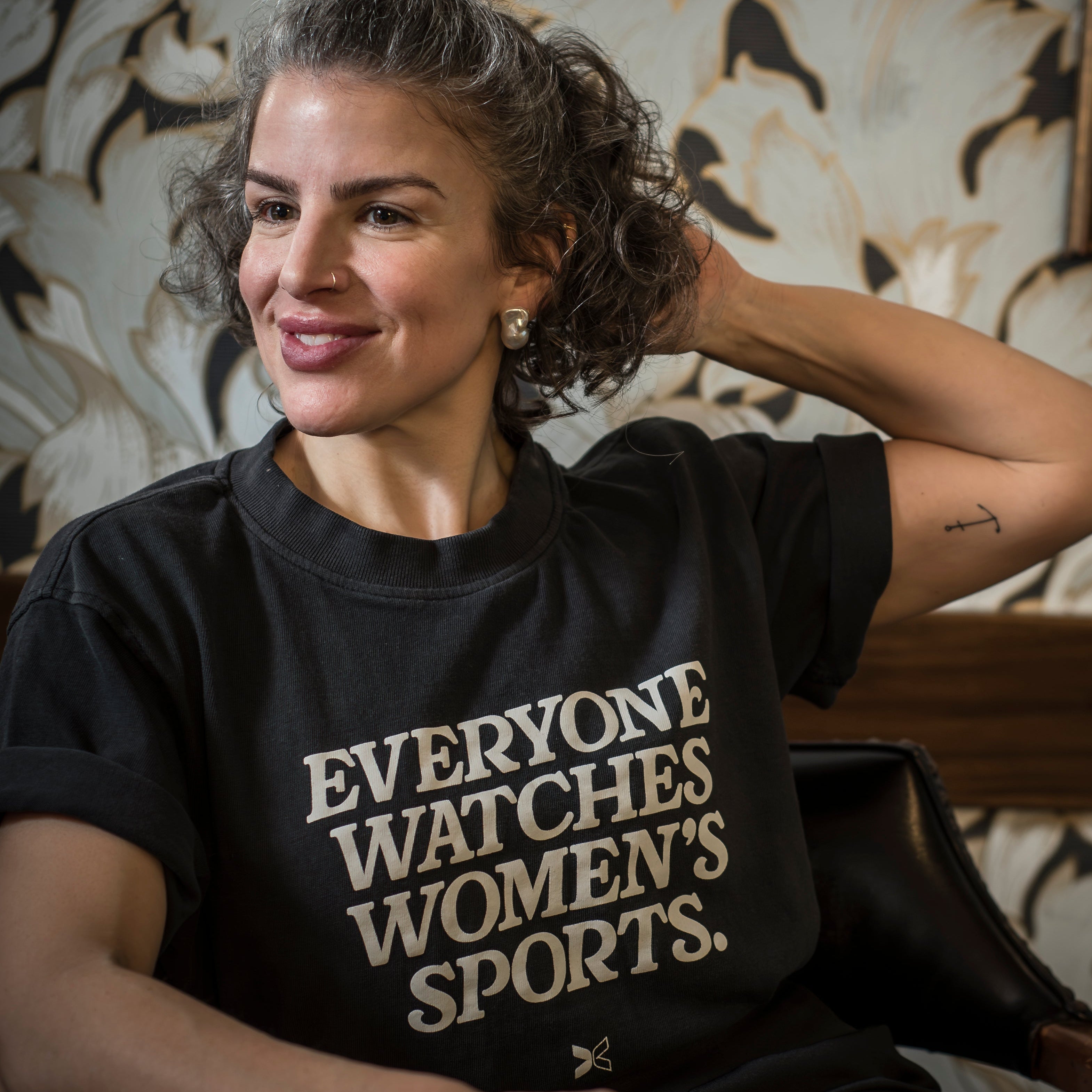 Woman wearing a black feminist t-shirt with text, wear large genuine baroque pearl stud earrings sitting in a stately steakhouse restaurant with vintage wallpaper.


