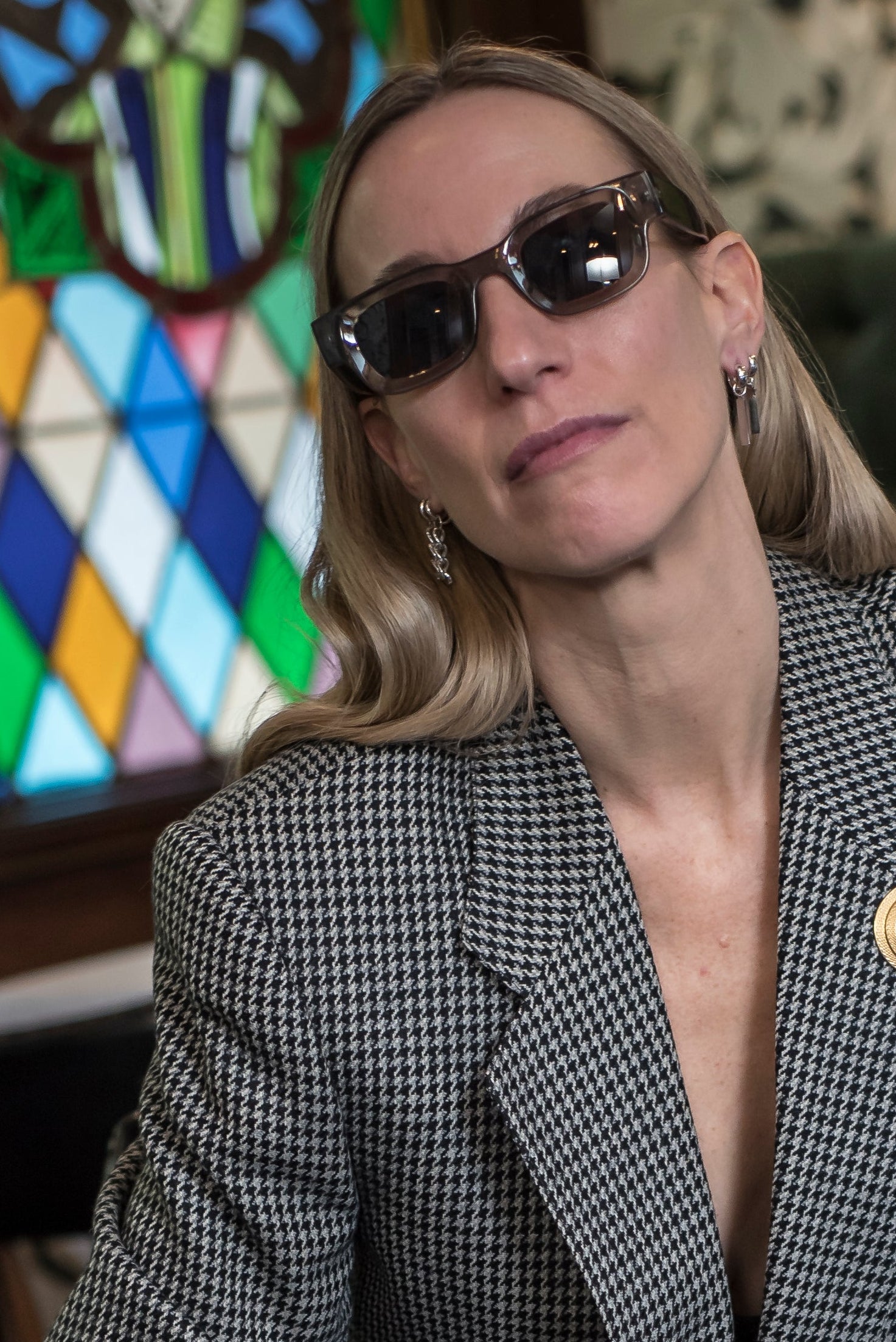 Woman sitting on a chair wearing sunglasses and a patterned blazer in a room with stained glass.